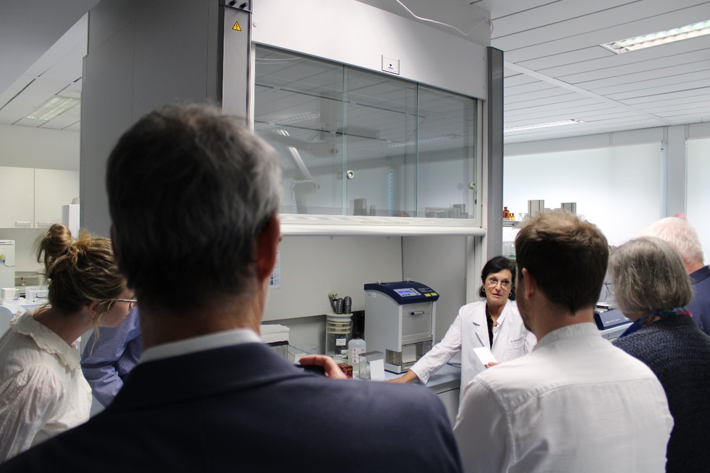 Das Bild zeigt eine technische Mitarbeiterin, die in einem Labor der Arzneimitteluntersuchungsstelle einer Besuchergruppe Analysenverfahren vorstellt.